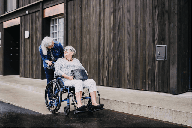 Elderly woman seated in STRONGBACK 24 Flip Wheelchair 1019 pushed by caregiver outdoors on wet pavement