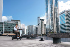 WHILL Model R Urban Mobility Scooter on a waterfront boardwalk with a city skyline in the background